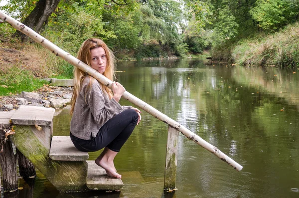 さまざまな感情の笑い、悲しみ、悲しみ、川や水を求めて森の銀行の木製の橋の心遣いで座って長いブロンドの髪を持つ美しい若い女性モデル — ストック写真