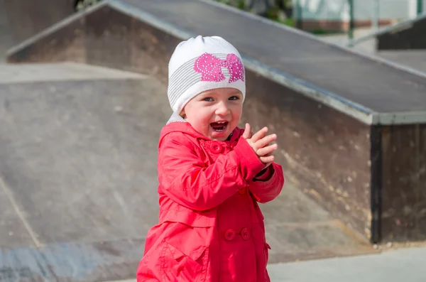 Bayi gadis cantik dengan jaket merah dan topi putih bermain di taman bermain skate, tersenyum dan bersenang-senang — Stok Foto