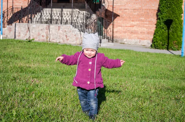 Pequeña niña hermosa abrigo de bebé, sombrero y jeans jugando en el parque caminando sobre hierba verde haciendo sus primeros pasos sonriendo y disfrutando de la felicidad — Foto de Stock