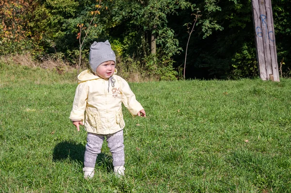 Pequeña niña hermosa abrigo de bebé, sombrero y jeans jugando en el parque caminando sobre hierba verde haciendo sus primeros pasos sonriendo y disfrutando de la felicidad — Foto de Stock