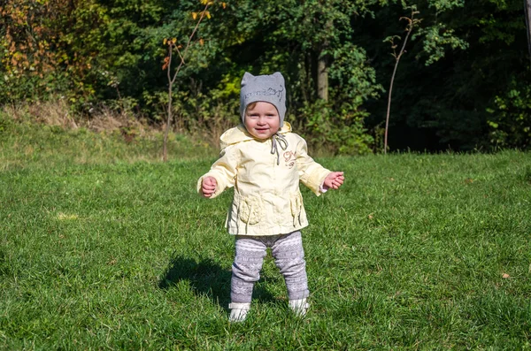 Bambina bella cappotto bambino, cappello e jeans che giocano nel parco camminando su erba verde facendo i loro primi passi sorridendo e godendo la felicità — Foto Stock