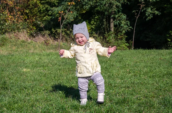 Bambina bella cappotto bambino, cappello e jeans che giocano nel parco camminando su erba verde facendo i loro primi passi sorridendo e godendo la felicità — Foto Stock