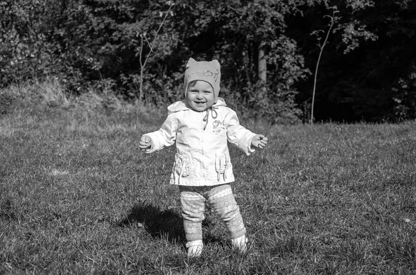 Mooi meisje baby jas, muts en jeans spelen in het park wandelen op groen gras doen hun eerste stappen glimlachend en genieten van geluk — Stockfoto