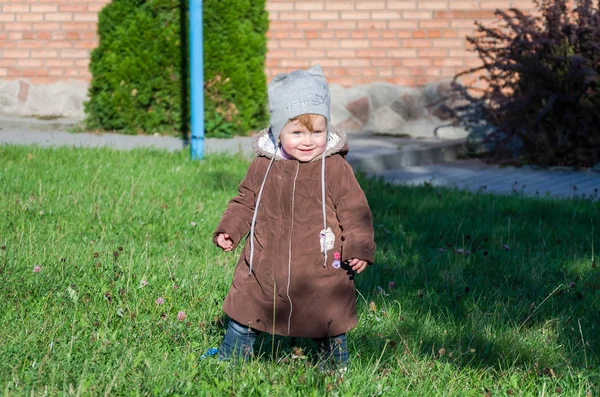 Gadis kecil yang cantik mantel bayi, topi dan jeans bermain di taman berjalan di rumput hijau melakukan langkah pertama mereka tersenyum dan menikmati kebahagiaan — Stok Foto
