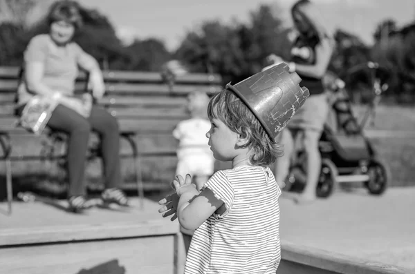 Маленькая очаровательная девочка играет на детской площадке в песочнице, на голове носит красное пластиковое ведро — стоковое фото