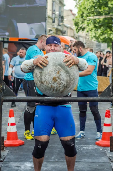 LVIV, UCRANIA - JUNIO 2016: Strong bodybuilder strongman levanta una enorme bola de piedra pesada hecha de mármol y la lanza sobre la barra en la calle bajo los ojos de los espectadores atónitos — Foto de Stock