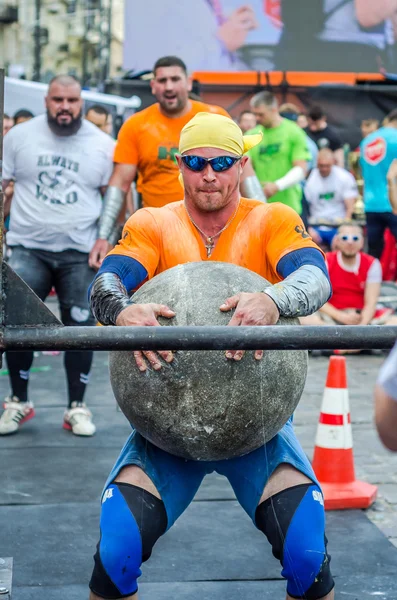 LVIV, UCRANIA - JUNIO 2016: Strong bodybuilder strongman levanta una enorme bola de piedra pesada hecha de mármol y la lanza sobre la barra en la calle bajo los ojos de los espectadores atónitos — Foto de Stock