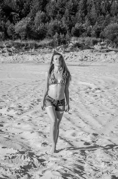 Young charming girl in short denim shorts and bikini swimsuit with long hair walking along the sand quarry sand extractive industrial scale