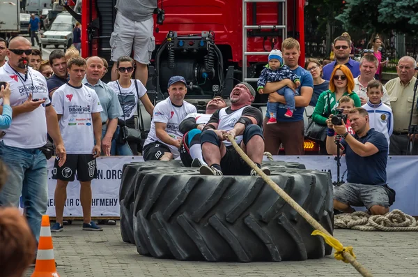 LVIV, UCRANIA - JULIO 2016: Fuerte atleta hombre fuerte tirando de un camión de cuerda sentado con un socio no es enorme neumáticos — Foto de Stock