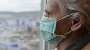 Tıbbi maskeli yaşlı kadın pencereden dışarı bakıyor.