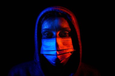 Siyah arka planda izole edilmiş kırmızı ve mavi ışıkla aydınlatılmış koruyucu bir tıbbi maske takan kapüşonlu genç bir kadının portresi.