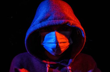 Siyah arka planda izole edilmiş kırmızı ve mavi ışıkla aydınlatılmış koruyucu bir tıbbi maske takan kapüşonlu genç bir kadının portresi.