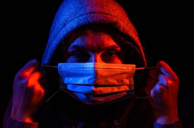 Siyah arka planda izole edilmiş kırmızı ve mavi ışıkla aydınlatılmış koruyucu bir tıbbi maske takan kapüşonlu genç bir kadının portresi.