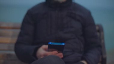 Tıbbi maskeli bir adam deniz kenarındaki bankta oturur ve telefona bakar.