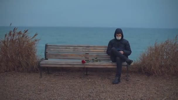 Загальний план. Чоловік у медичній масці сидить на лавці біля моря і дивиться на телефон — стокове відео