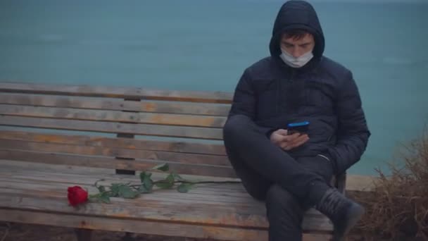 En man i medicinsk mask sitter på en bänk nära havet. — Stockvideo