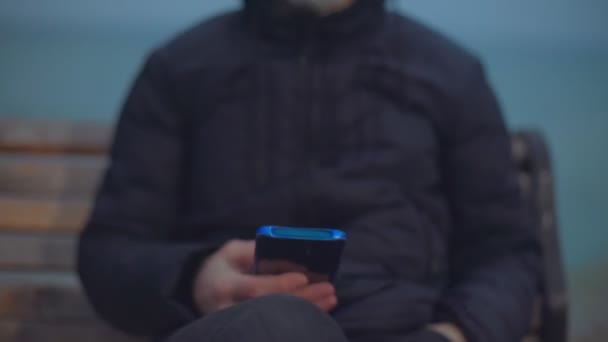 Чоловік у медичній масці сидить на лавці біля моря і дивиться на телефон — стокове відео