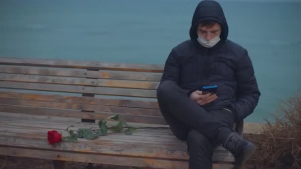Мужчина в медицинской маске сидит на скамейке у моря и смотрит в телефон — стоковое видео