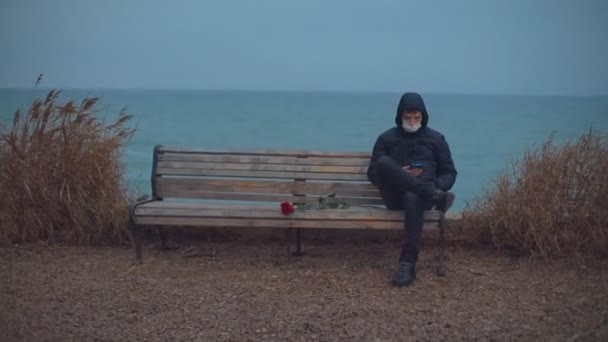 Загальний план. Чоловік у медичній масці сидить на лавці біля моря і дивиться на телефон — стокове відео