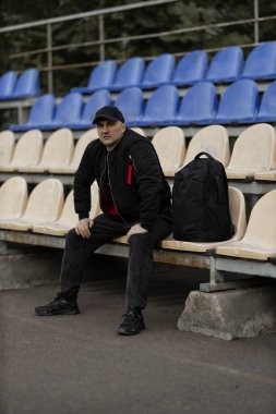 man dressed in a black bomber jacket and cap sits quietly in an empty stadium tribune. He has a backpack beside him and appears to be enjoying a moment of solitude.