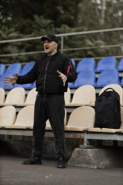 In a quiet stadium a man in a black bomber jacket and cap stands in the empty stands expressing strong emotions as he reacts to the game unfolding. His energy adds life to the scene.