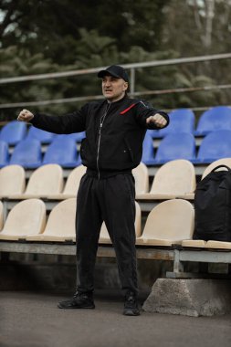 man stands in empty stadium wearing a black bomber jacket and cap expressing excitement and support for his team as game progresses on field nearby.