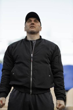 man in black bomber jacket and cap stands in empty stadium showing reactions to the team performance during an intense game feeling excitement and tension.
