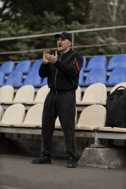man stands in the empty stadium wearing a black bomber jacket and cap. He claps and reacts enthusiastically to the teams game showing his support for the players.