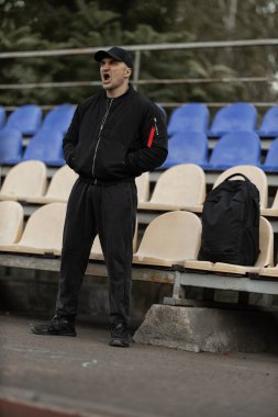 man dressed in a black bomber jacket and cap stands alone in the empty stands showing his emotions as he watches the game unfold. He is focused and engaged with the action on the field.