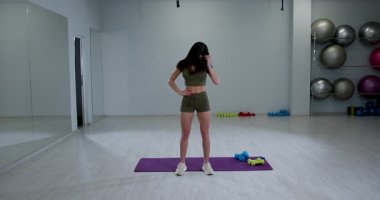 Atletik kadın, esnekliğini arttırmak için kafa döndürme egzersizi yaparken minderin üzerinde duruyor. İyi aydınlatılmış bir fitness stüdyosunda, etrafta egzersiz ekipmanları var..