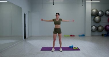 Spor kıyafeti giymiş atletik bir kadın, parlak bir spor salonunda kol halkaları çiziyor. Alan, arka planda görünür egzersiz ekipmanları ve ağırlıklarla geniştir..