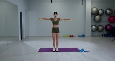 Atletik kadın, temiz, parlak bir spor salonunda yükselen halterlerle meşgul. Vücudunun üst kısımlarına kararlı bir şekilde odaklanarak güçlü bir duruş sergiliyor..