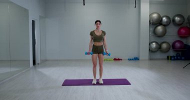 Atletik kadın, kendine güvenen bir spor salonunda ayakta duran halterli biseps kıvrımlarına odaklanıyor. Hareketlerini arttırmak için uygun bir kıyafet giyiyor..