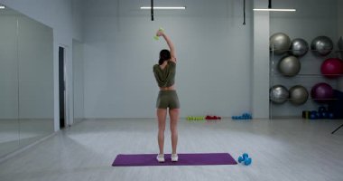 Atletik kadın, bir kolunu kullanarak başının üstüne dambıl kaldırırken sırtı kameraya dönük durur. Odaklanmış ve spor salonu spor aletleri ve spor aksesuarlarıyla dolu..