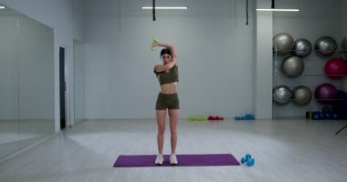 Atletik kadın, geniş bir fitness stüdyosunda tek kollu halterli triseps uzantısını gösteriyor. Parlak ışıklandırma, gücünü ve dayanıklılığını hedeflediği için odaklanmış eğitim rutinini geliştirir..