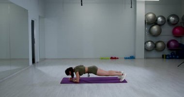 Atletik kadın, iyi aydınlatılmış bir spor salonunda yoga minderinde ön kol tahtası kullanıyor. Ayarlar egzersiz ekipmanları ve aynaları içeriyor. Fitness için motive edici bir atmosfer yaratıyorlar..
