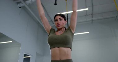 Atletik kadın iyi aydınlatılmış bir spor salonunda dururken kol halkaları gösteriyor. Gücünü ve esnekliğini arttırma amaçlı bir sabah egzersizi sırasında kaslarını çalıştırarak vücuduna odaklanır..