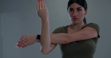 Atletik kadın, iyi aydınlatılmış bir spor salonunda omuz kaslarını esnetiyor. Formuna ve tekniğine odaklanmış vaziyette. Akşamları spor rutinine bağlılığını gösteriyor..