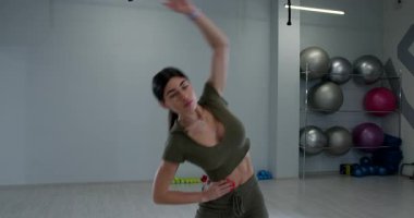Atletik kadın modern bir spor salonunda ayakta esneme egzersizi yapıyor. Parlak geniş bir çalışma ortamında denge ve esnekliği korumaya odaklanıyor..