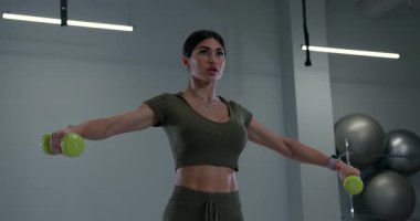 Atletik kadın spor salonundaki antrenmanına odaklanıyor. Dambıl kaldırarak omuz kaslarını güçlendiriyor..