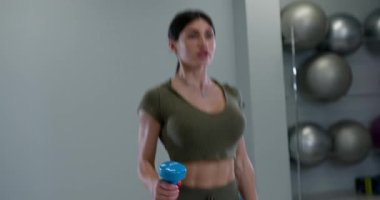 Atletik kadın tek kollu bir halter bisepsi ile meşgul. Mavi bir dambılı kaldırırken şekline odaklanıyor. Spor, spor aletleriyle dolu modern bir spor salonunda yapılıyor..