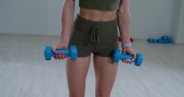 Atletik bir kadın, parlak bir fitness stüdyosunda iki dirhem bir çekirdekte duruyor. Vücuduna ve gücüne odaklanan açık mavi bir çift dambıl tutuyor..