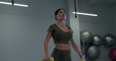 Formda bir kadın, parlak geniş bir spor salonunda yanal halter kaldırır. Atmosfer enerji dolu ve egzersiz oturumları için ideal..