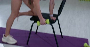 Atletik bir kadın destek için sandalye kullanırken tek kollu bir halterle egzersiz yapıyor. İyi ışıklandırılmış bir spor salonunda egzersiz rutinine odaklanmış durumda..