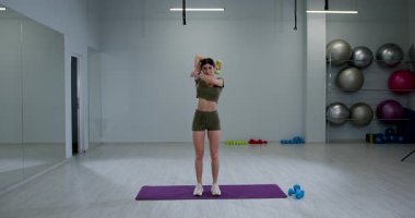 Atletik kadın modern bir spor salonunda tek kollu halterli trisepsi çalıştırıyor. Egzersiz aletleri ve ağırlıklarla çevriliyken formuna odaklanıyor..