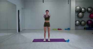 Atletik kadın iyi aydınlatılmış bir spor salonunda grup çalışması yapıyor. Yoga minderinin üzerinde dururken direnç bandı kullanarak vücudunun üst kısmını güçlendirmeye odaklanmış..