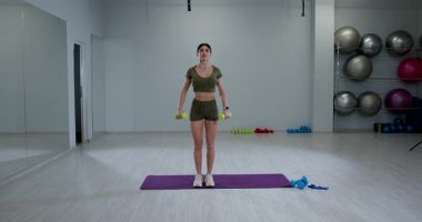 Atletik kadın, iyi aydınlatılmış bir spor salonunda yanal halterler yükseltiyor. Antrenman kıyafetleri giyiyor ve antrenman sırasında ağırlık kaldırırken odaklanmış bir ifade takınıyor..