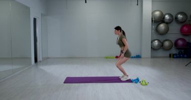 Atletik kadın, iyi aydınlatılmış bir spor salonundaki spor minderinin üzerinde diz şınav çekiyor. Yakınlarda çeşitli ağırlıklar görülebilirken şekline odaklanıyor. Bu eğitim oturumu dayanıklılığı ve uygunluğu arttırıyor..