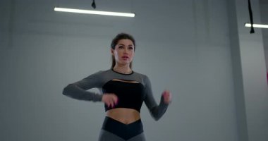 Atletik bir kadın, iyi aydınlatılmış bir spor salonunda ayakta duruyor. Aktif giyimde güçlü bir duruş sergilerken esnekliğine odaklanıyor..