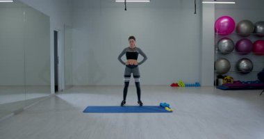 Sporcu bir kadın spor salonundaki mavi bir paspasın üzerinde duruyor. Spor aletleri ve büyük egzersiz toplarıyla çevrili antrenmanına odaklanırken başını öne ve arkaya eğiyor..